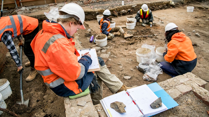 Five people in high-vis clothing and hard hats digging and cataloguing items on an excavation site.