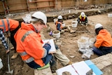 Five people in high-vis clothing and hard hats digging and cataloguing items on an excavation site.