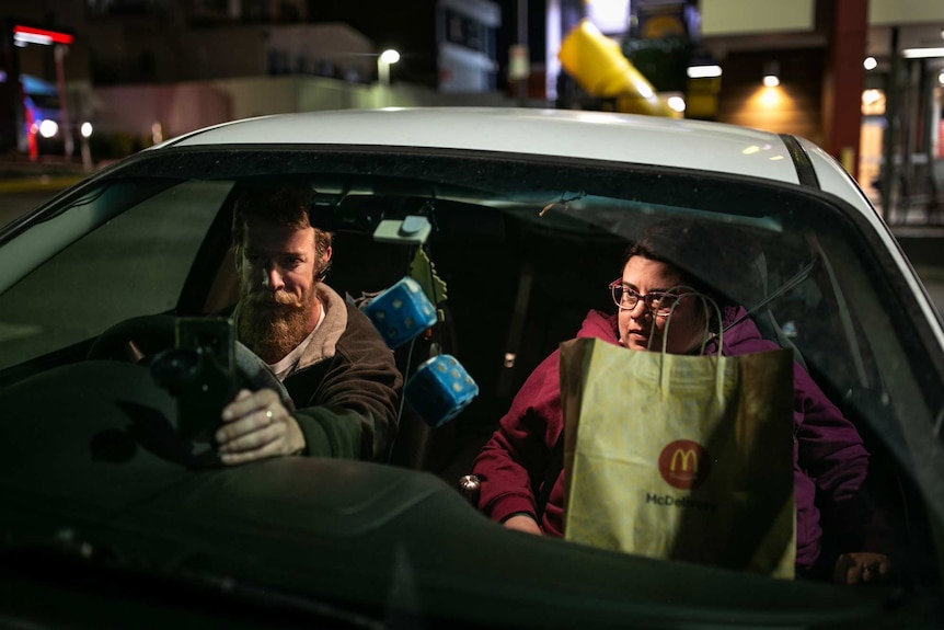A man and a woman sit in a car holding. He touches a mobile phone while she holds McDonald's packaging.