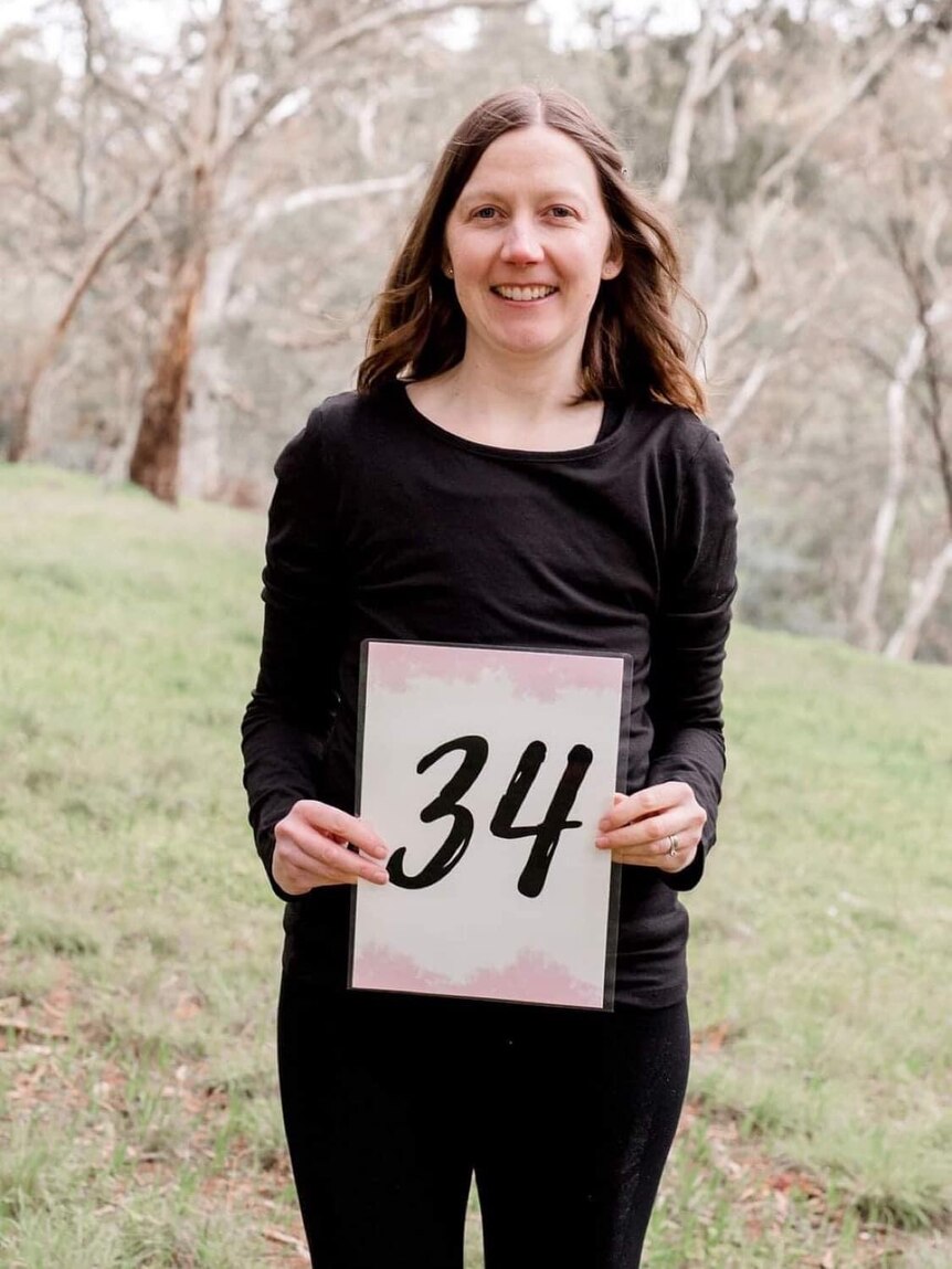 Catherine King smiles while holding up a sign with the number 34, her age when she was diagnosed with breast cancer.