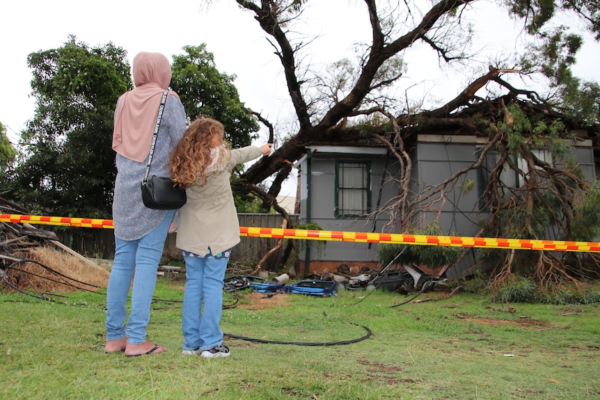 Child and woman stand next to safety tape looking at house that tree has fallen on