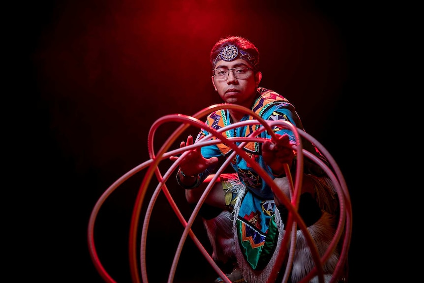A man in bright blue Native American regalia poses with spherical shape created from hoops in front of red and black background.