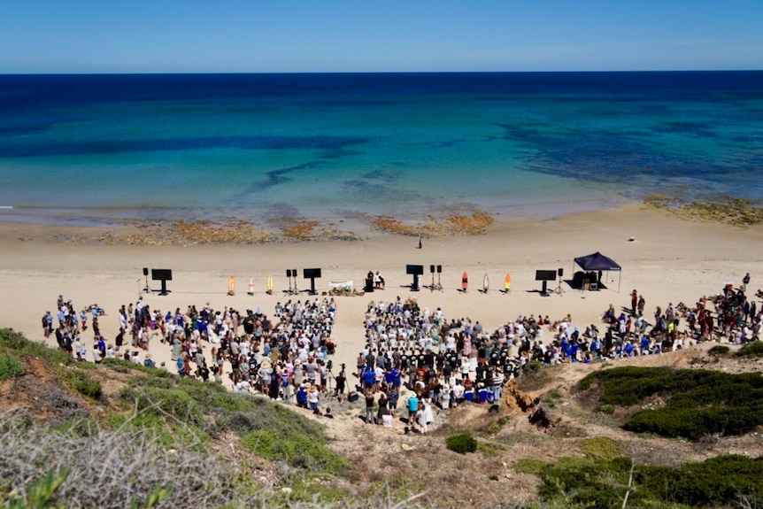 Wide shot from above of people gathered at a funeral service on a beach. 