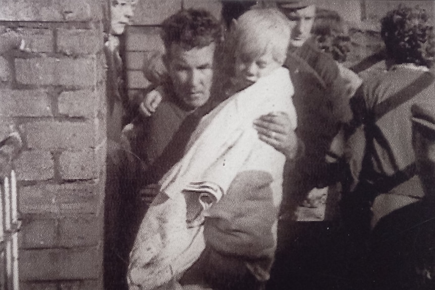 A small blonde-haired boy wrapped in a blanket is carried by a man.