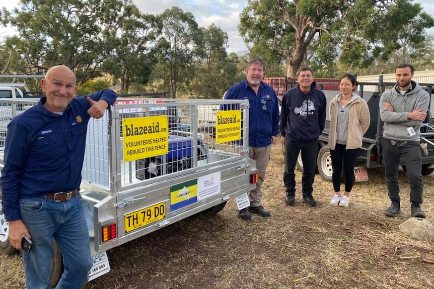 four men and one woman stand next to a trailer on a farm