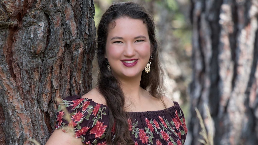 A young woman wearing gold earrings stands next to a tree in a floral dress, smiling.