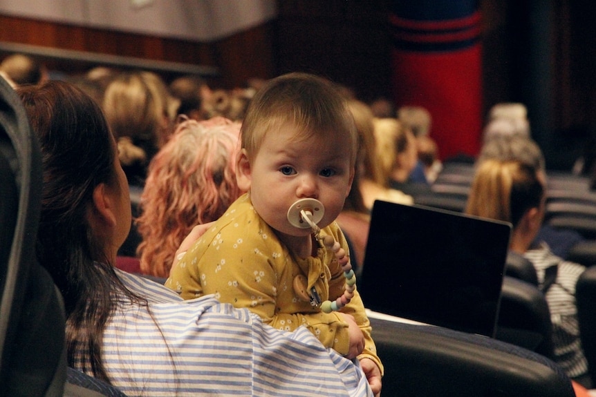 small baby looks at camera over mums shoulder in lecture theatre