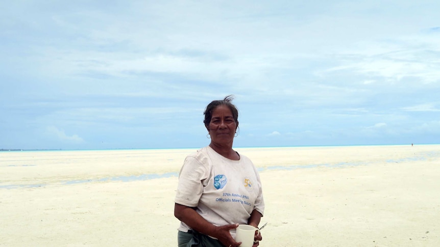 A woman standing on a sandy flat with the ocean in the distance. She is holding a white mug.
