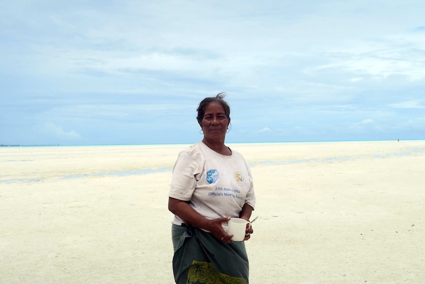 A woman standing on a sandy flat with the ocean in the distance. She is holding a white mug.