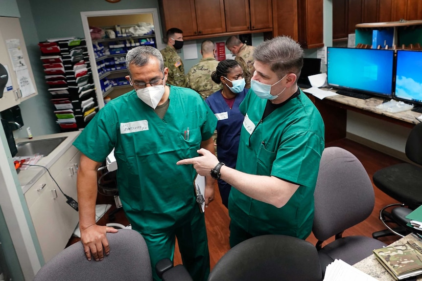 Two men wearing green scrubs and face masks speak in the middle of a medical office.