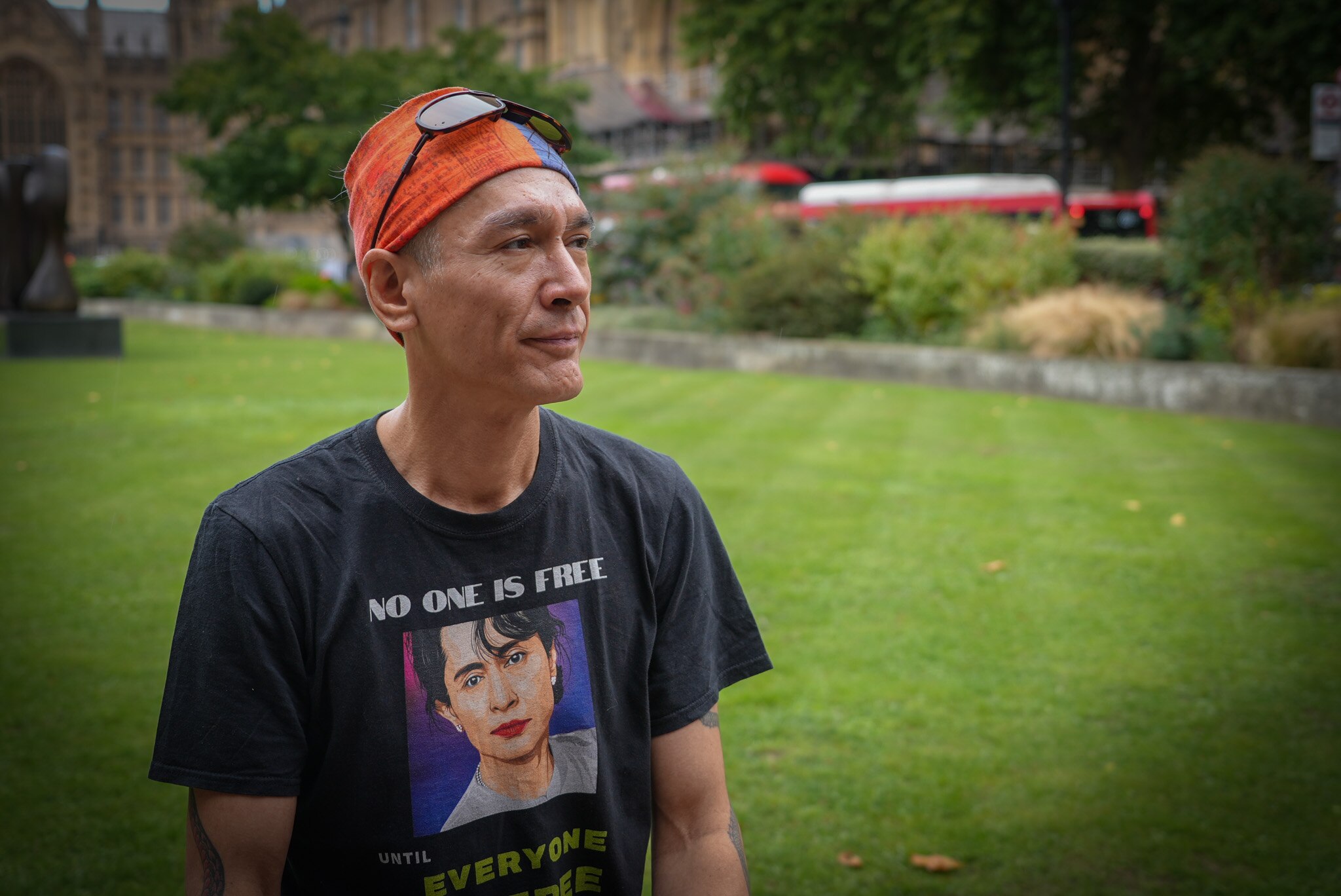 A man wearing sunglasses over an orange bandana stands in a park.