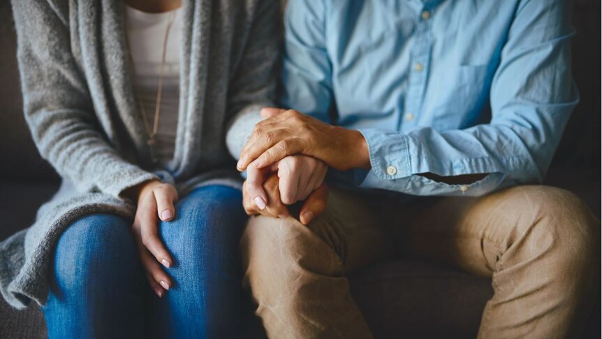 The slightly blurred bodies of a woman and a man, seated, with their hands held and resting on the man's knee.