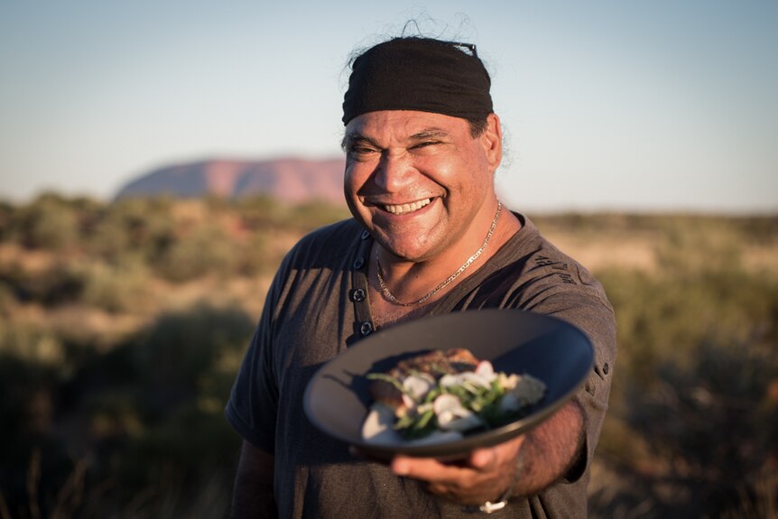 Mark Olive stands in front of Uluru holding a plate of bush tucker
