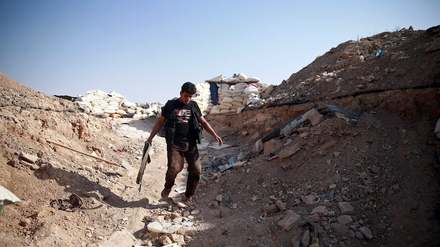 A man holding a gun in one hand walks down a hill between sandbags.