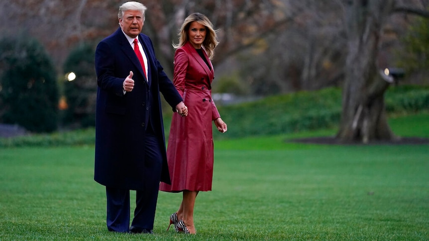 Donald Trump and Melania Trump holding hands while standing on a grass lawn. She is in a red coat.