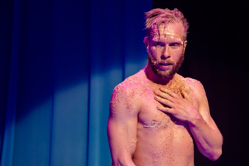 Head and torso shot of young man with short light hair and beard, bare but skin and face covered in colourful candy.