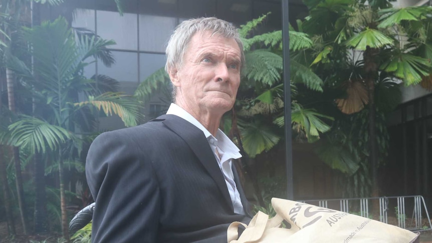 Man with grey hair wears a suit in front of a court house while sitting in a wheelchair with a serious, tired expression on his