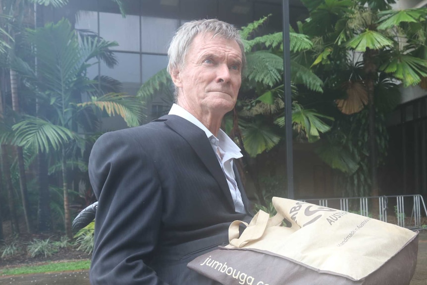 Man with grey hair wears a suit in front of a court house while sitting in a wheelchair with a serious, tired expression