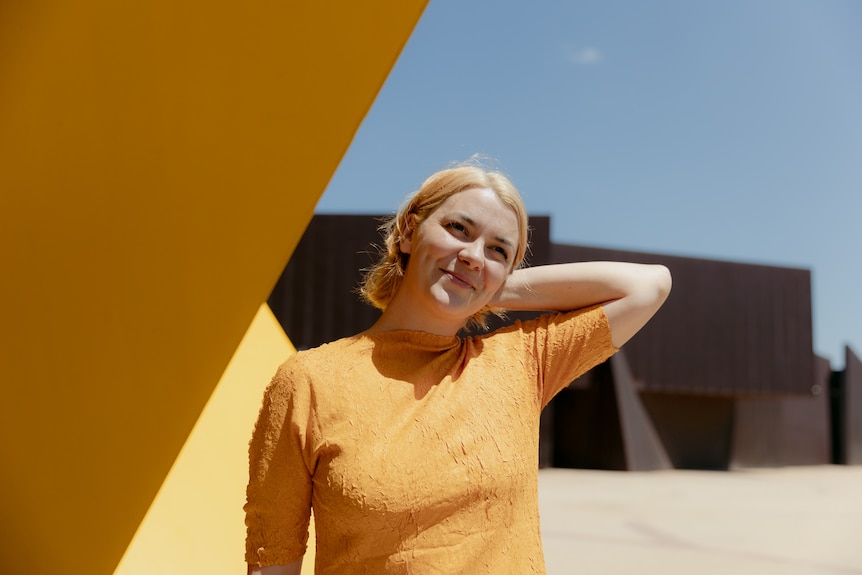A Wiradjuri woman with light blonde hair and brown eyes wears a yellow crepe dress and stands beside a bright yellow building.