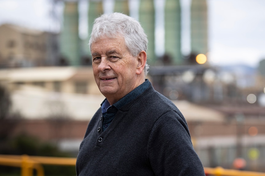 An grey-haired man stands in front of a factory.