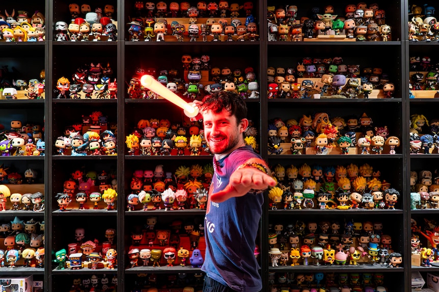 Man holds glowing light saber up in front of shelves of collectible pop vinyls. 