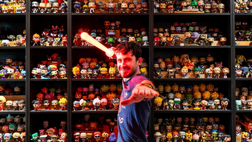 Man holds glowing light saber up in front of shelves of collectible pop vinyls. 