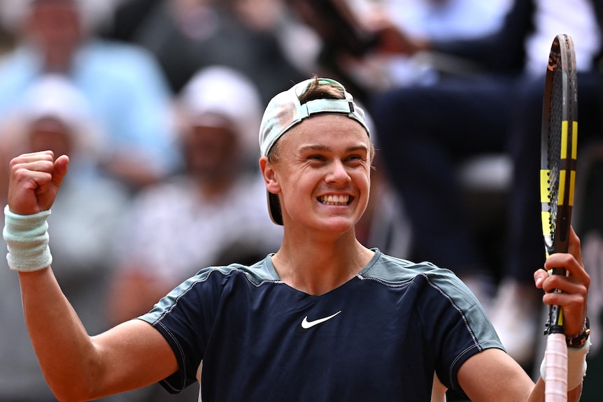 A tennis player with his cap backwards smiles as he holds his arms aloft.
