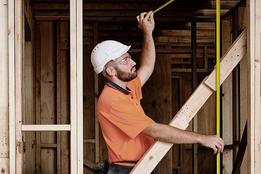 a young  man wearing a hard hat while holding a measuring tape standing in a construction site