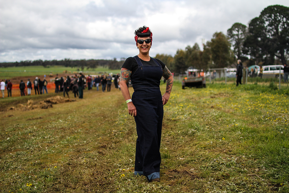 A woman with a 50s hairdo, tattoos and denim overalls stands in a grassy paddock.