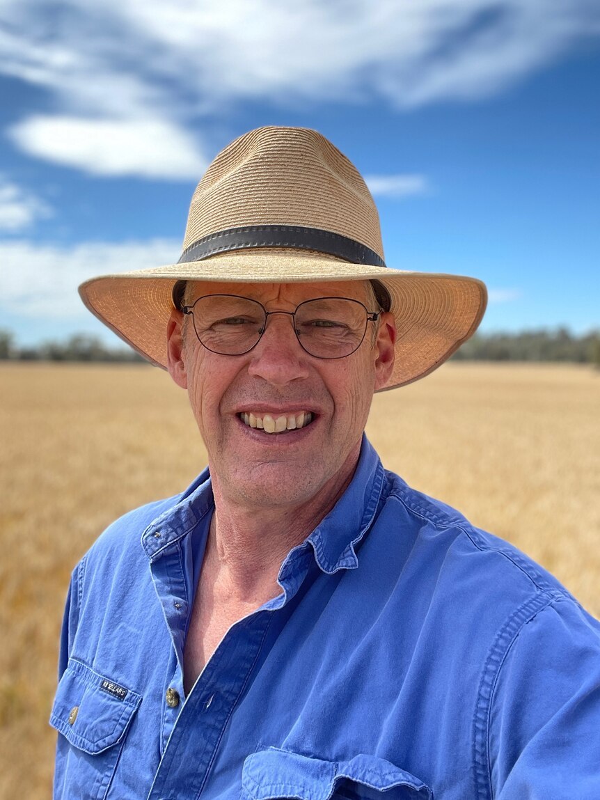 A man with a blue shirt and straw hat smiles at the camera in a wheat crop