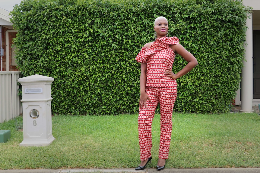 Woman standing out the front of her home. She's wearing a bright red top and pants.