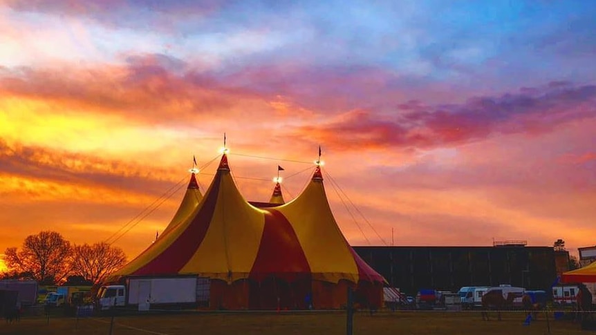 Under a pink and blue sunset, a large red and yellow coloured circus tent stands, with trucks in front of it.
