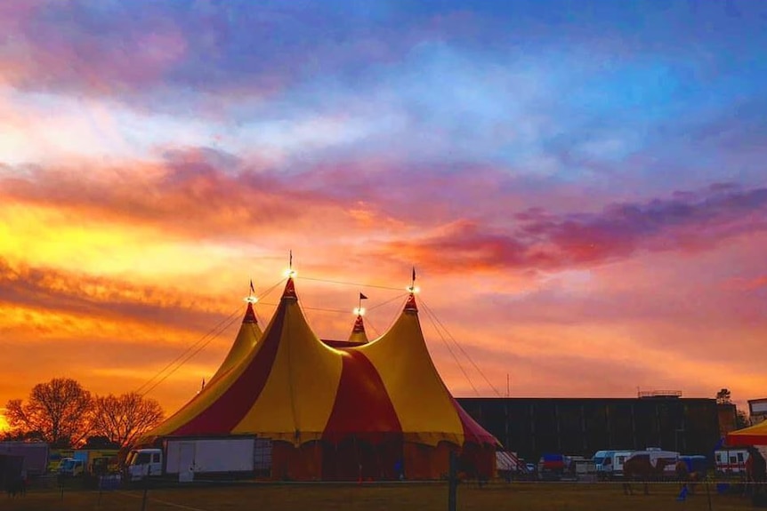 Under a pink and blue sunset, a large red and yellow coloured circus tent stands, with trucks in front of it.