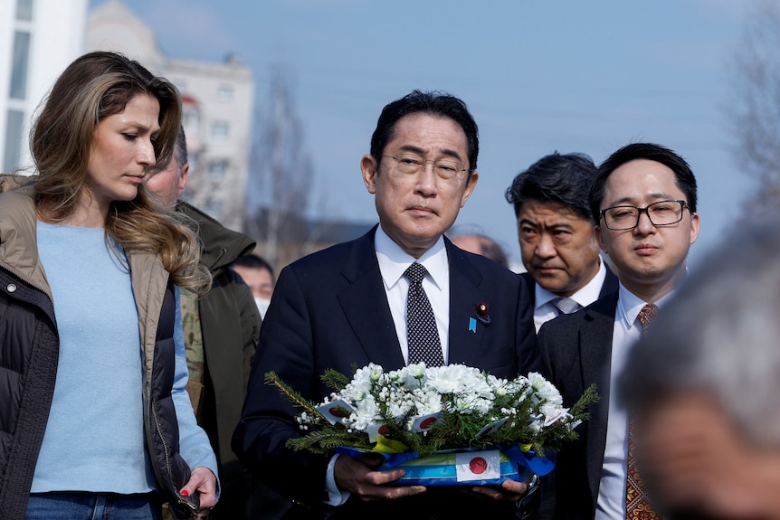 Kishida holds a wreath of white flowers on a yellow and blue base stamped with the Japanese flag