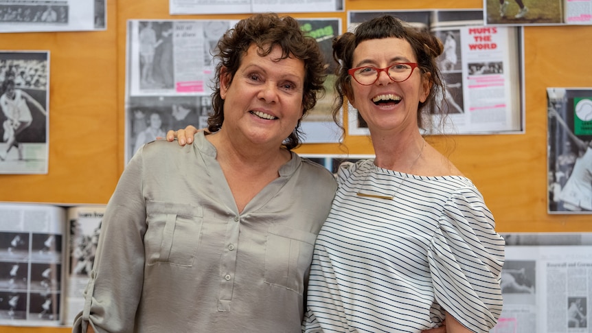 Two women stand with their arms around each other, smiling widely. In the background are newspaper cuttings of Evonne's career