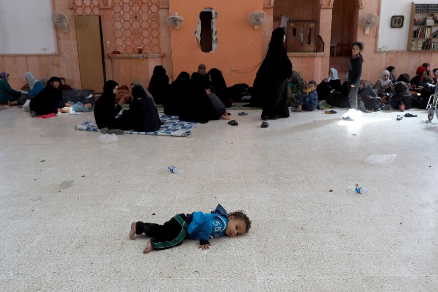 A child lies on the cold floor of the mosque as women share food and talk behind him.