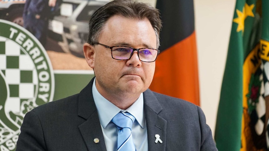 A man in glasses stands in front of flags at a press conference.