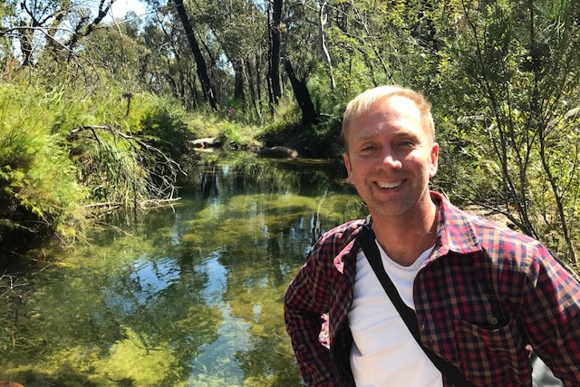 A smiling man with short hair standing in front of a bush creek.