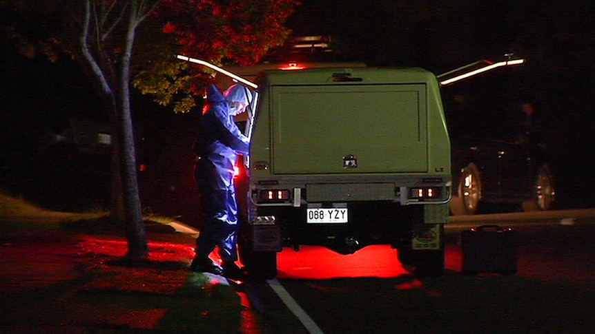 Man in blue forensic suit and hair net stands at back of car at night