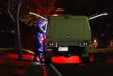 Man in blue forensic suit and hair net stands at back of car at night