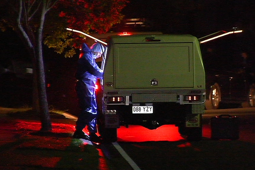 Man in blue forensic suit and hair net stands at back of car at night