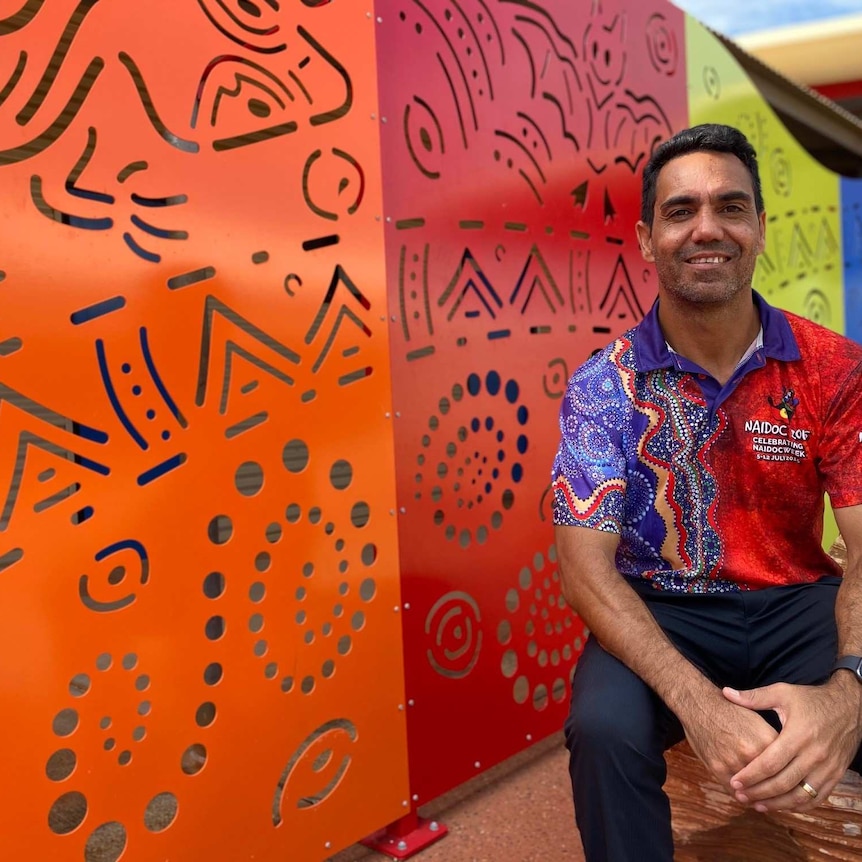 A man sits on a rock in front of bright coloured laser cut sheets showing indigenous designs