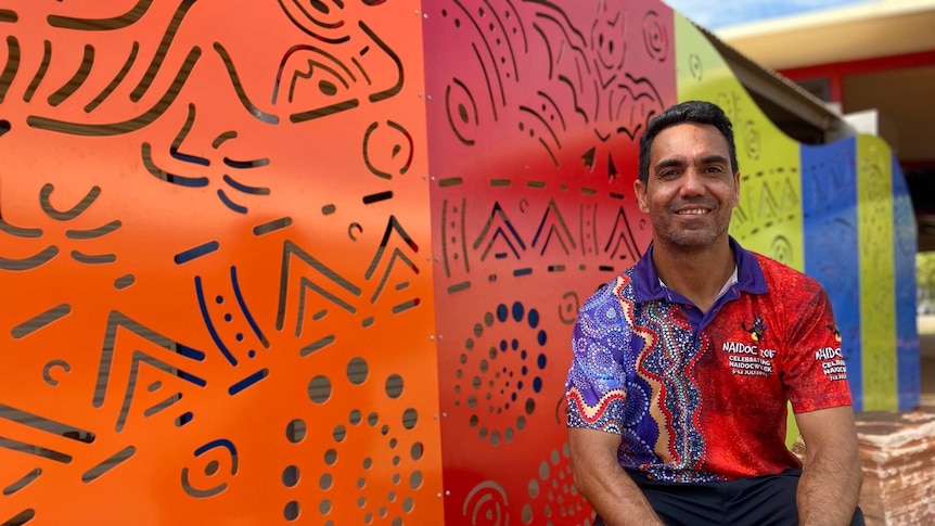 A man sits on a rock in front of bright coloured laser cut sheets showing indigenous designs