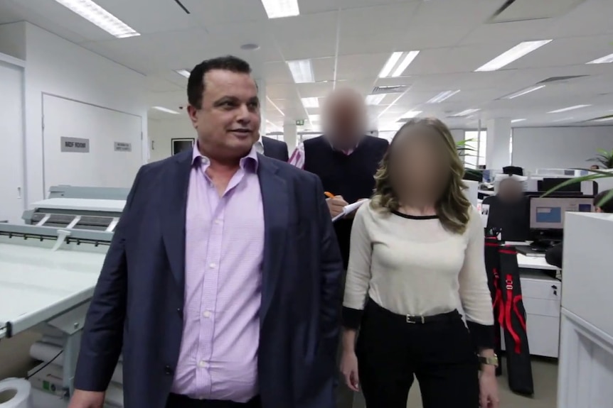 A man wearing a suit jacket leads three people walking through an open plan office.