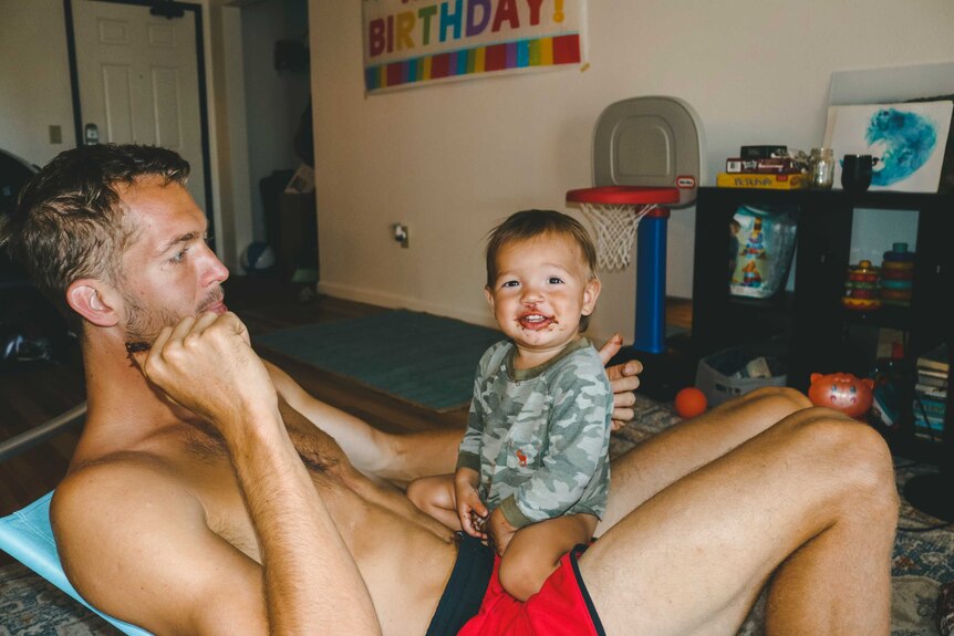 A man wearing just shorts in a loungeroom with a baby with a messy face on his lap