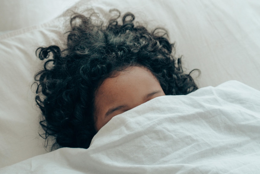 A person with brown curly hair in bed with the sheets pulled up to their eyes