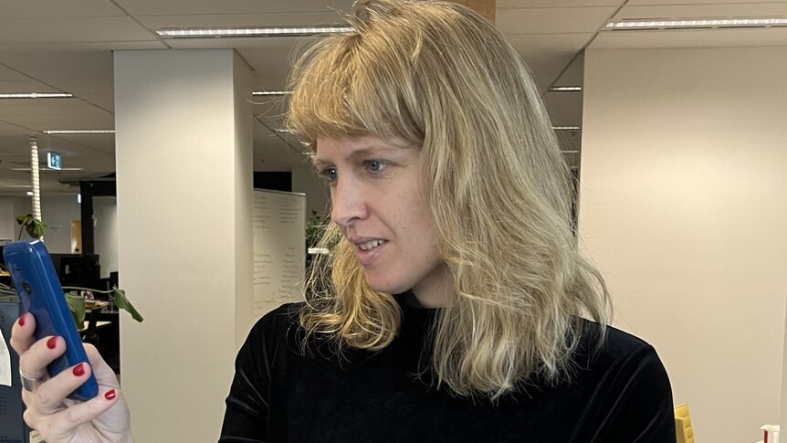 Rosa Ellen, with black shirt and blue pants, and shoulder-length blonde hair, stands in an office looking at a phone screen.