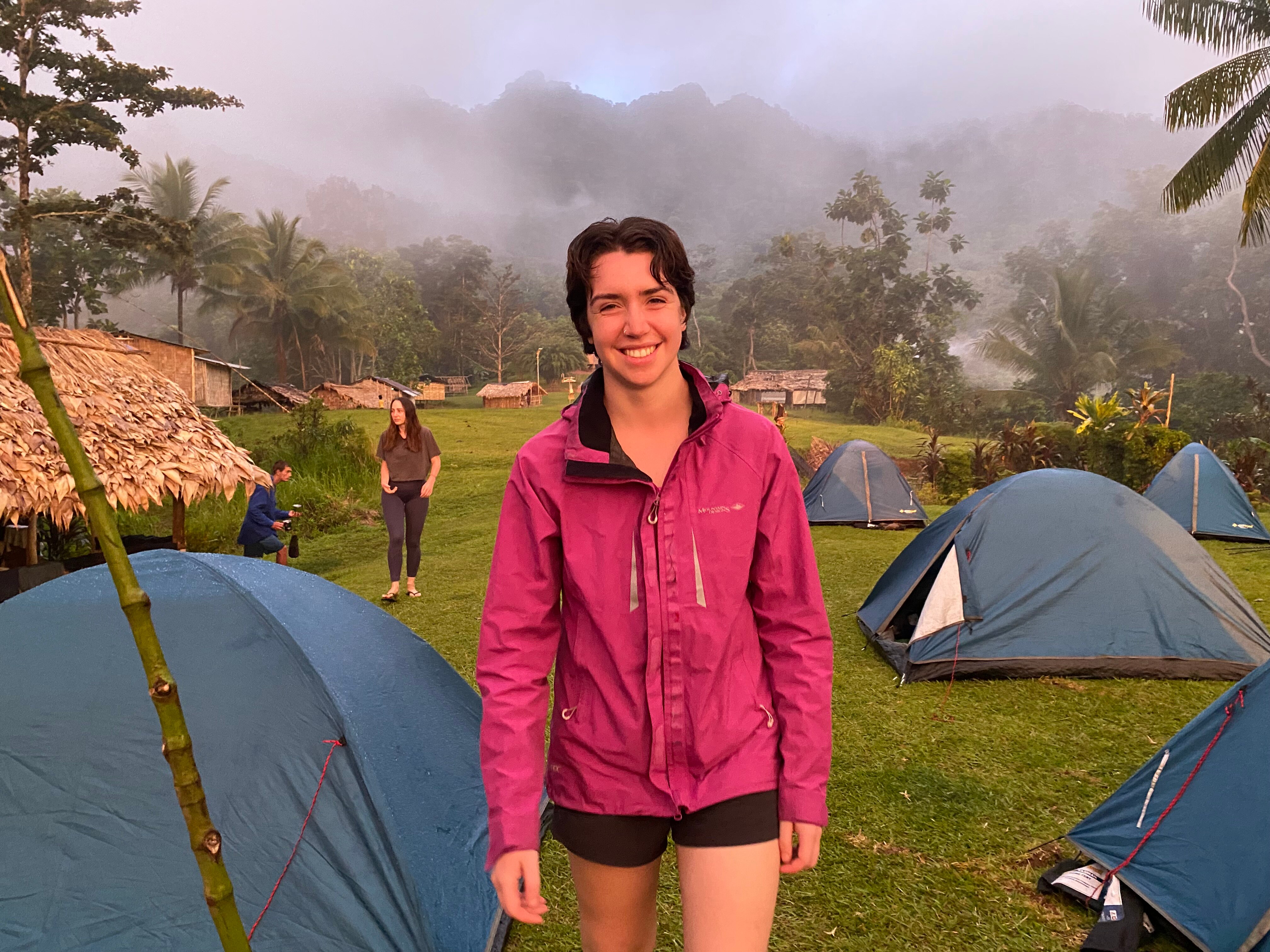 A young woman wearing hiking gear stands in a misty field amongst small tents. 