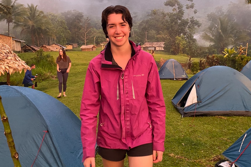 A young woman wearing hiking gear stands in a misty field amongst small tents. 
