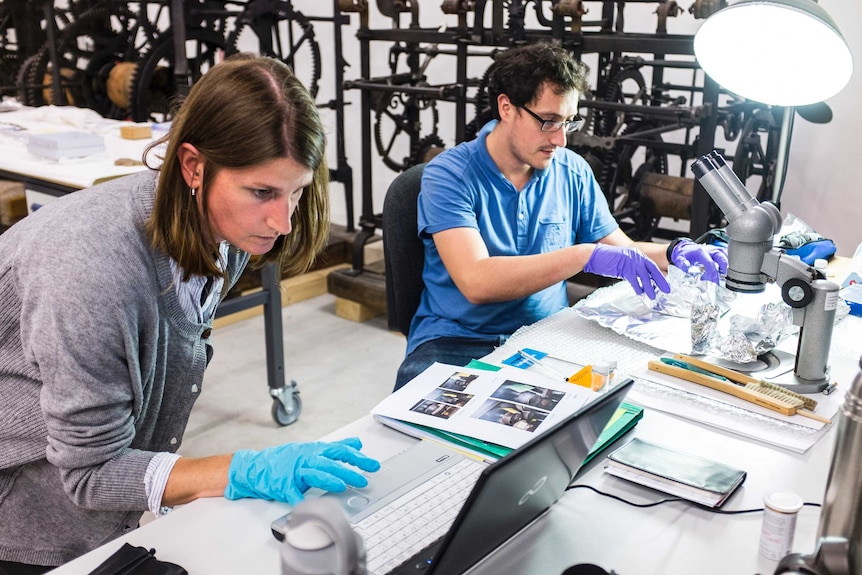 Two scientists work at a desk, one is handling a sample near a microscope, the other in on a laptop